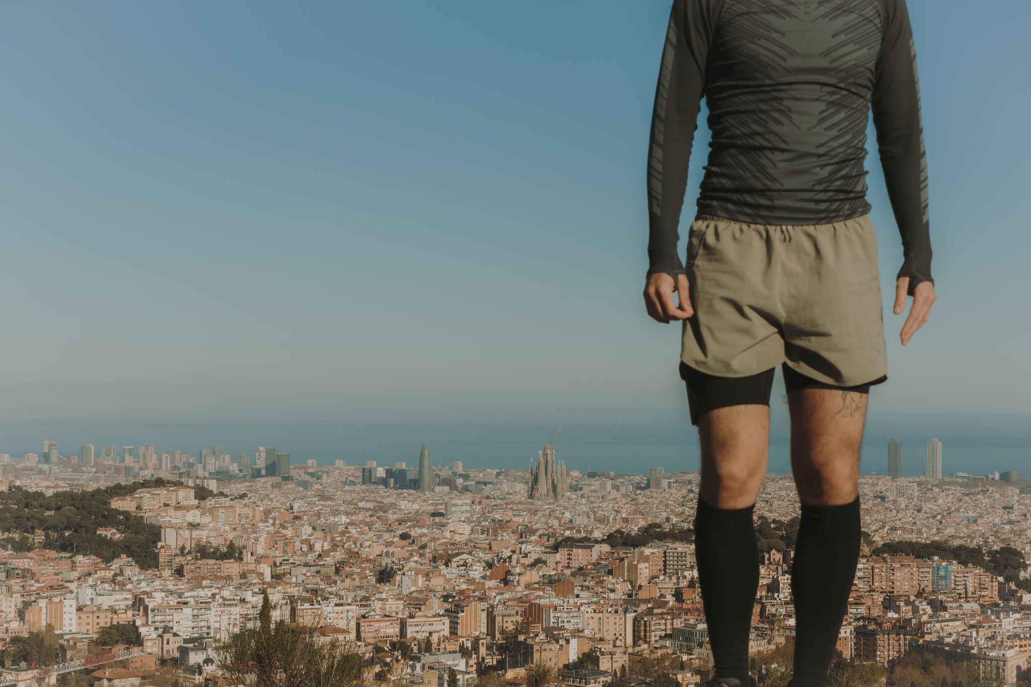 Man standing in an elevated mountain area, with a panoramic view of Barcelona, the Sagrada Família and the sea in the background.