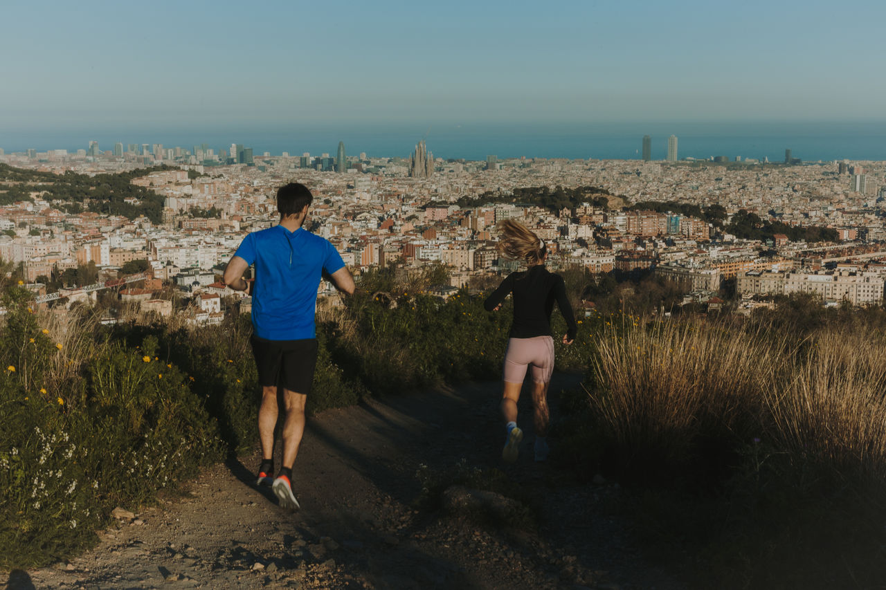 Dues persones corrent per un camí de terra amb una vista panoràmica de Barcelona, la Sagrada Família i el mar al fons.