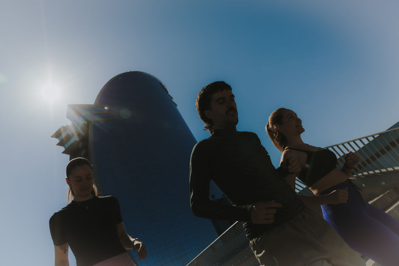 Tres persones corren amb l’Hotel W Barcelona al fons, sota un cel blau i amb el sol a contrallum
