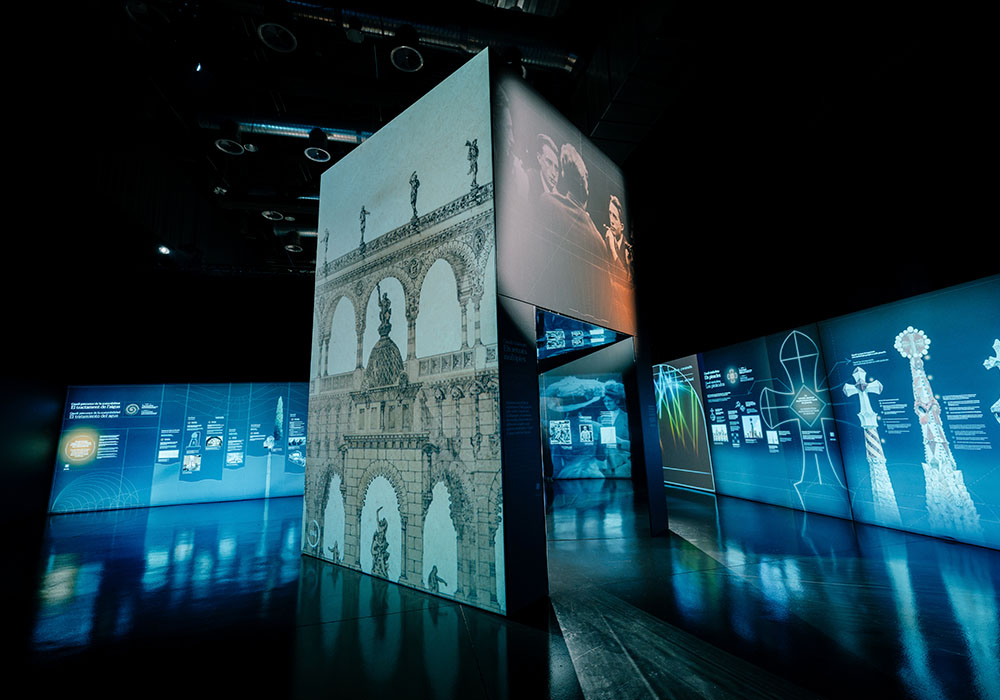 Immersive exhibition space with architectural projections and visual content on large screens, with reflections on the floor.