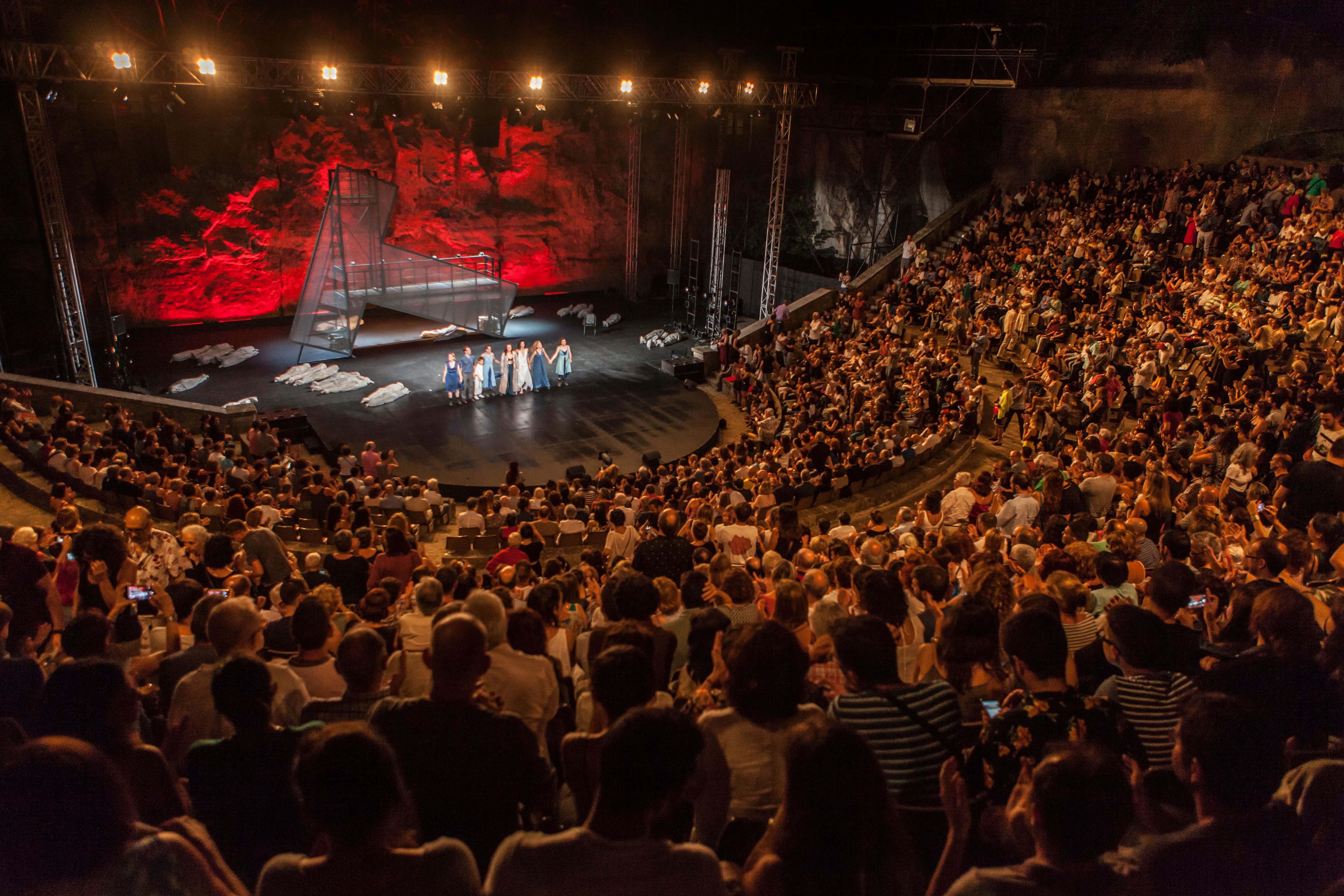 Large open-air theater filled with an audience seated in semicircular stands. On the central stage, several actresses are taking a bow while figures lie on the floor covered with light-colored fabrics. The background of the stage is illuminated with intense red lights against a rocky wall.