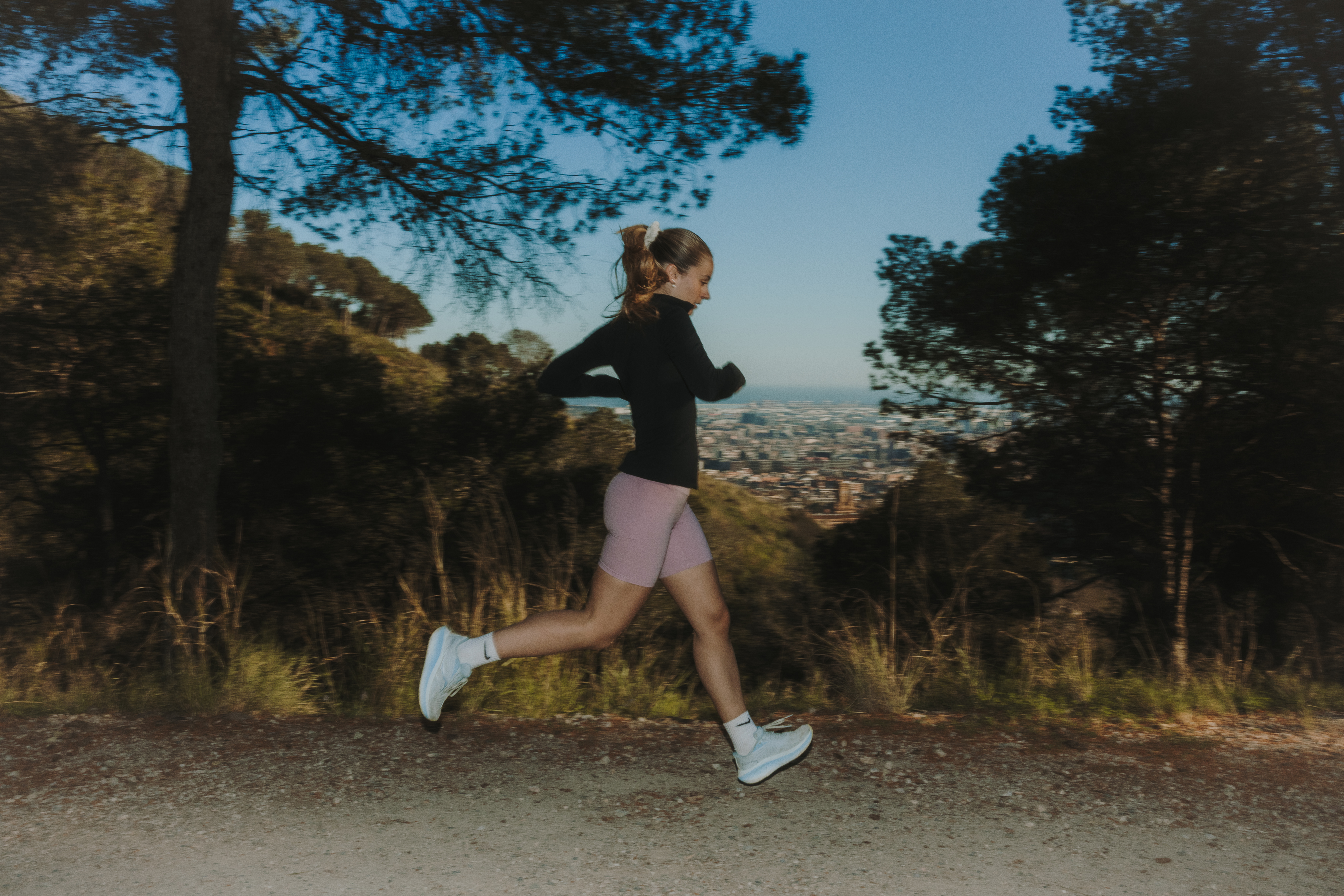 Girl running along a dirt path among trees, with a panoramic view of Barcelona in the background.