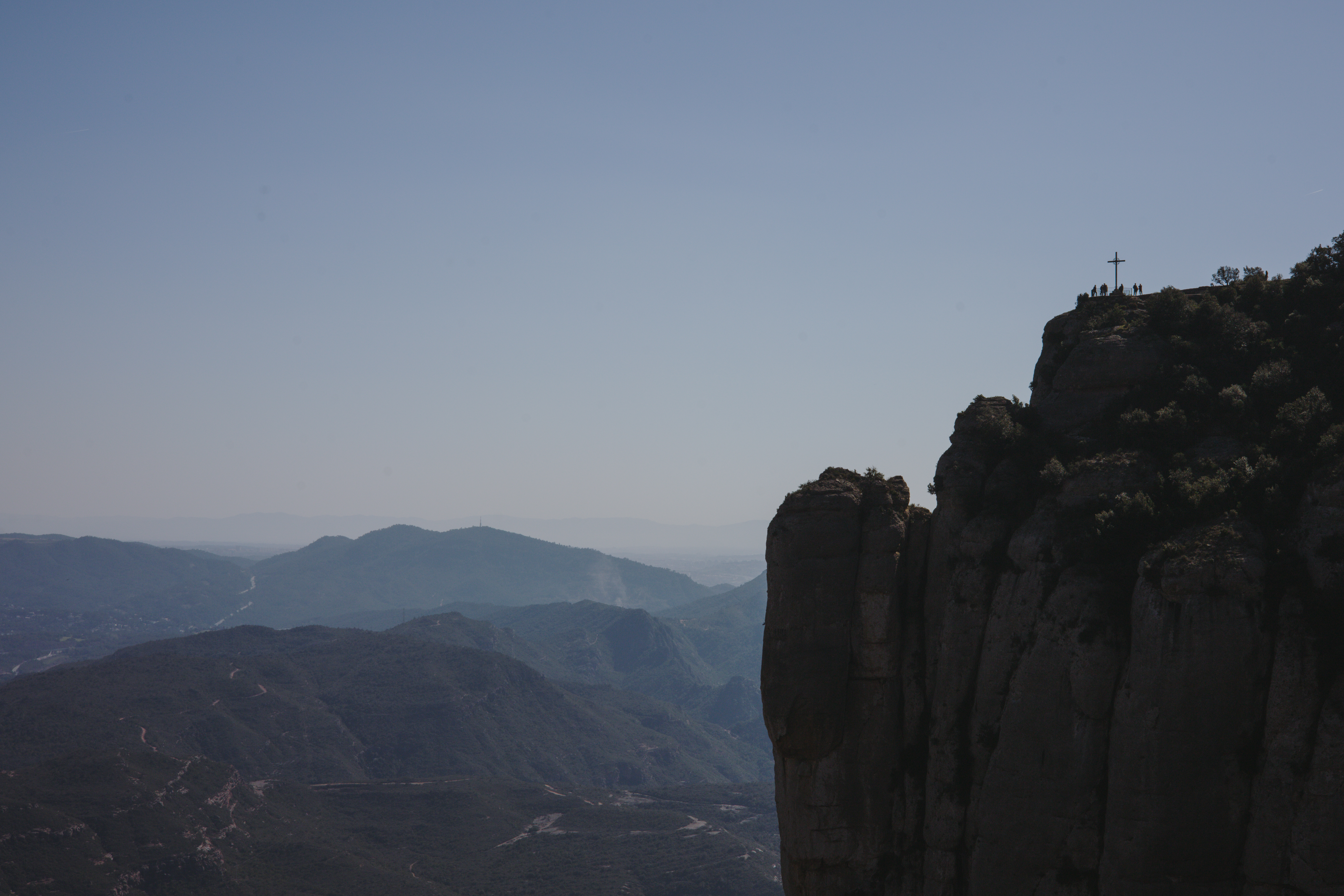 Cliff of Montserrat with the Sant Miquel cross at the summit, with green landscape at the foot of the mountain.