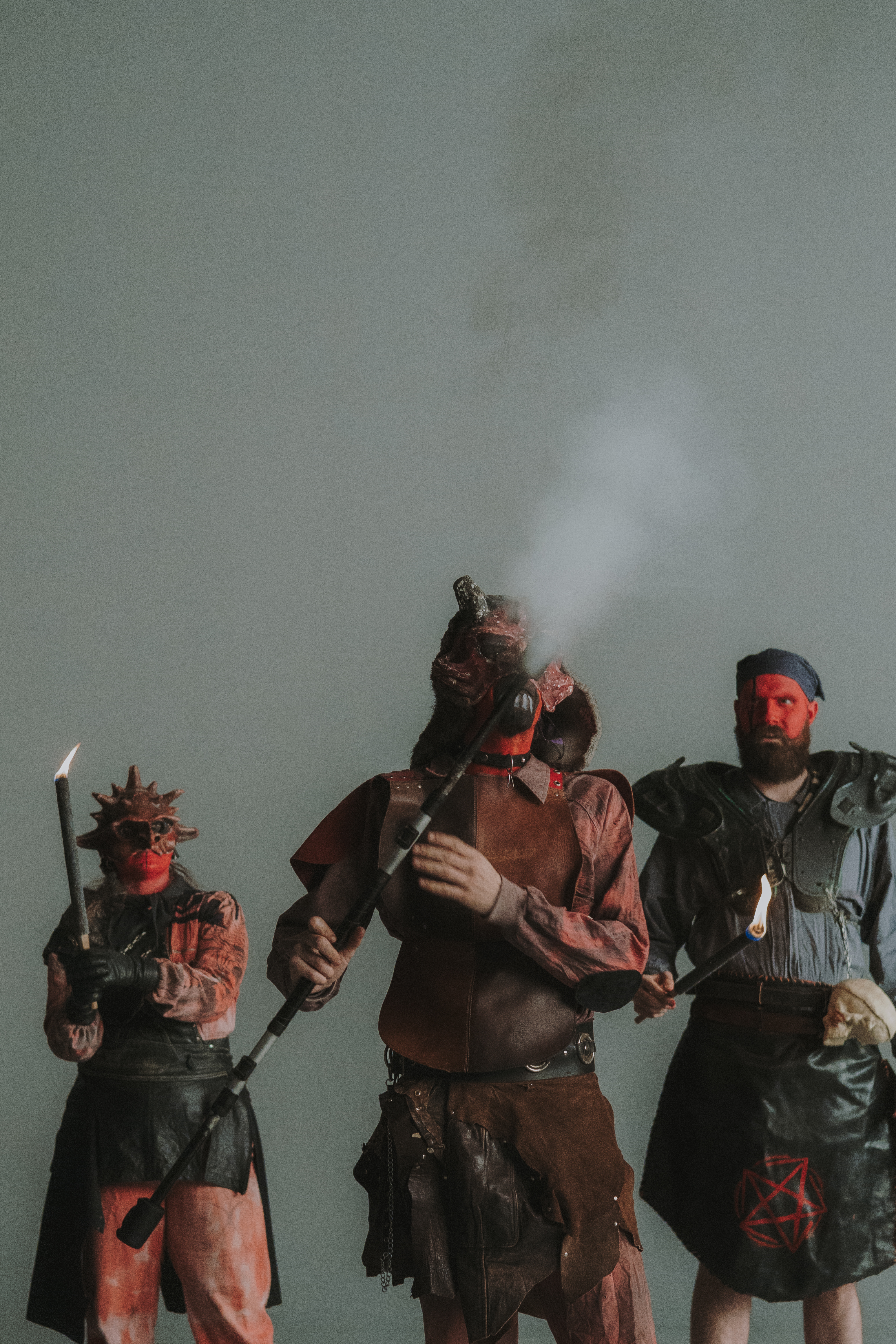 Tres personas vestidas con disfraces de diablos sostienen antorchas y un bastón humeante, con rostros pintados o enmascarados y vestuario protector. La escena, sobre un fondo neutro, evoca la tradición del correfoc y el mundo festivo del fuego.