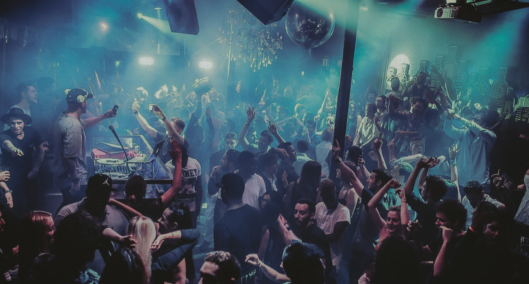 Nightclub filled with people dancing under blue lights and smoke, with a DJ performing in an elevated booth while the crowd raises their hands or records with their phones, creating a festive and vibrant nighttime atmosphere.