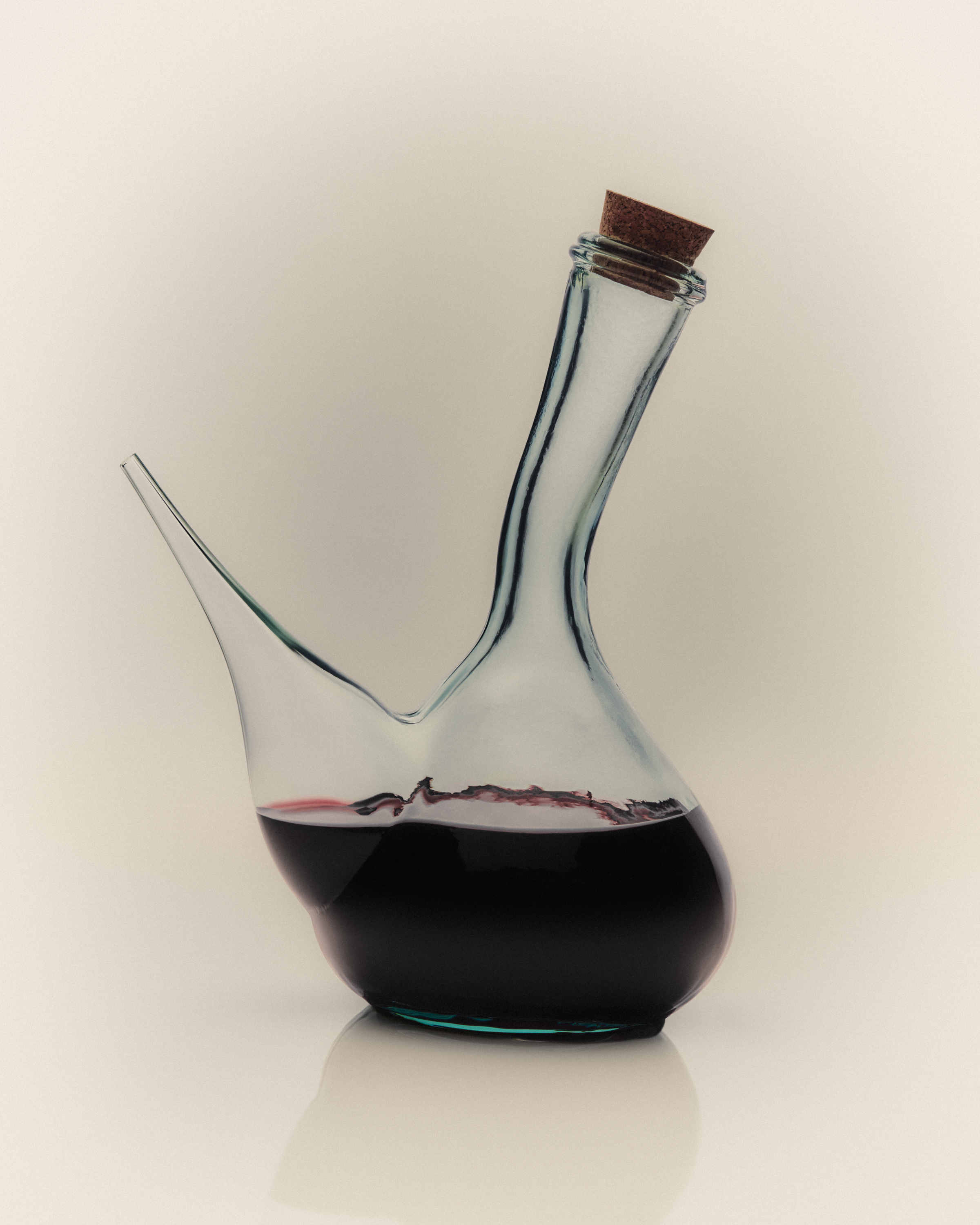 Transparent glass wine decanter with a long, angled neck, partially filled with red wine, against a light neutral background.