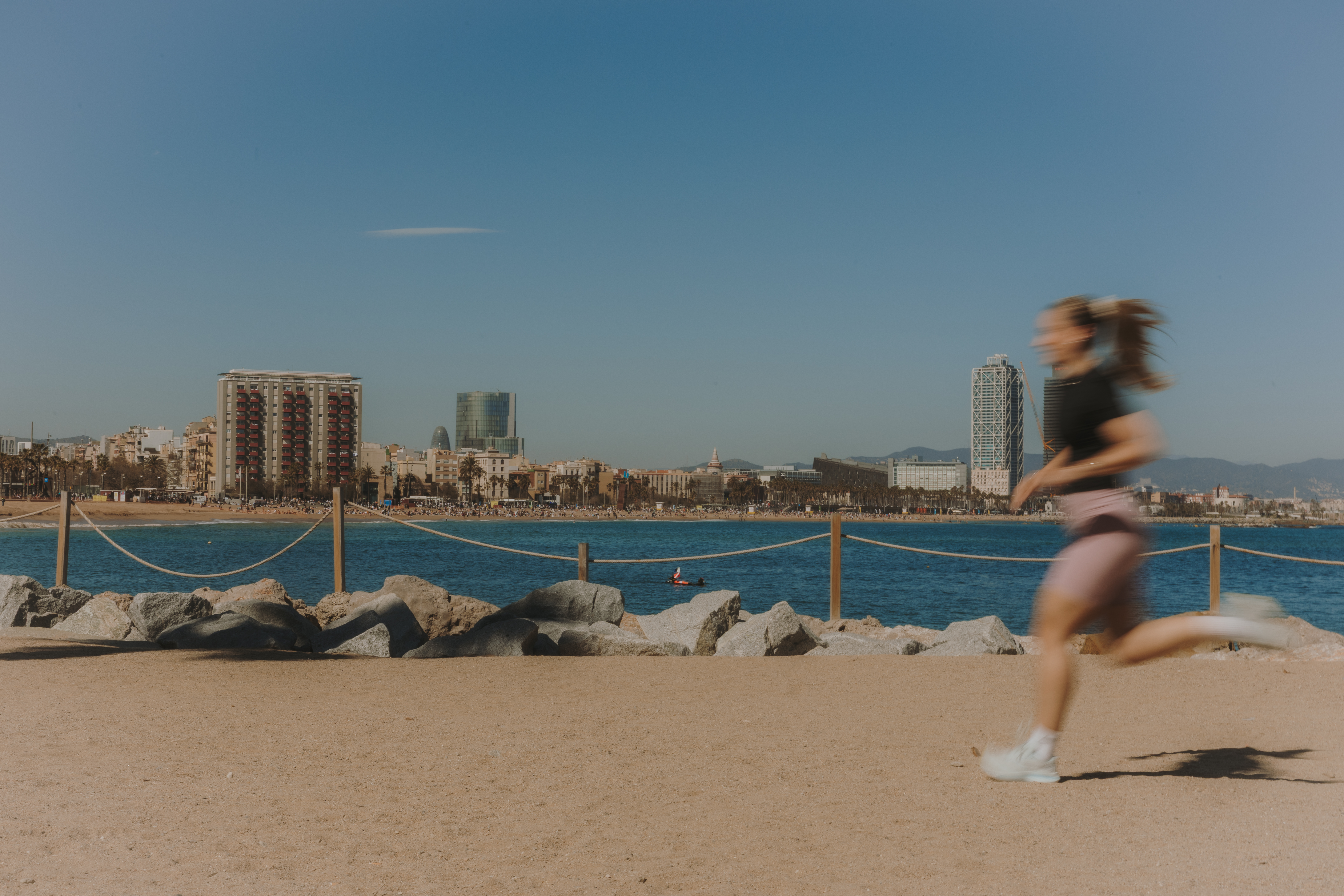 Girl running along the seafront promenade. In the background, several buildings of the Barcelona skyline under a cloudless blue sky on a sunny day.