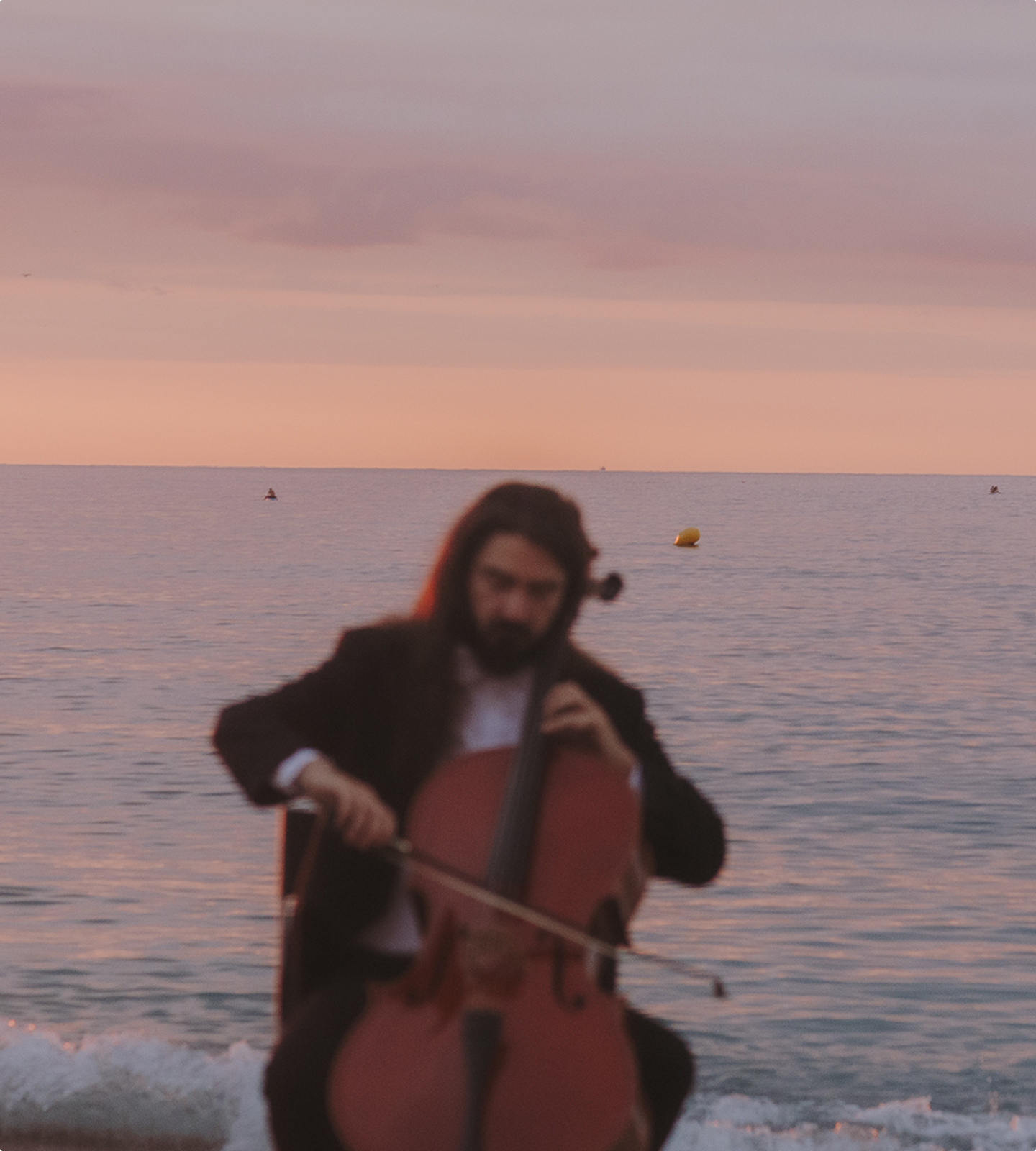 A bearded man in formal attire plays the cello on the beach, with the sea in the background and a sunset sky.
