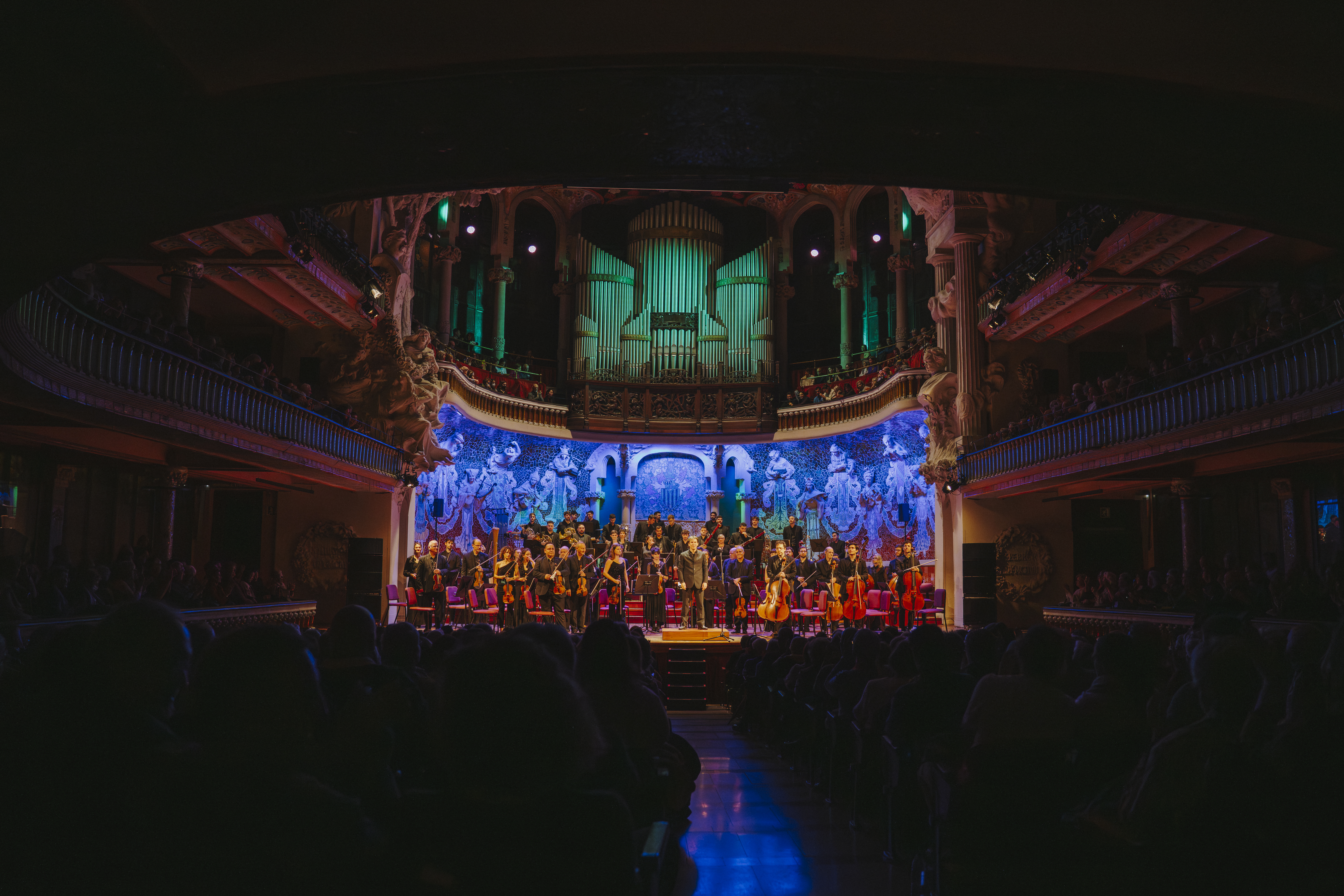 A concert hall with a large organ at the back and a stage full of musicians and singers during a performance. The space is majestic, with side balconies, sculptural decoration, and an illuminated mural behind the orchestra. The audience appears in the foreground, seated in the dark while watching the show.