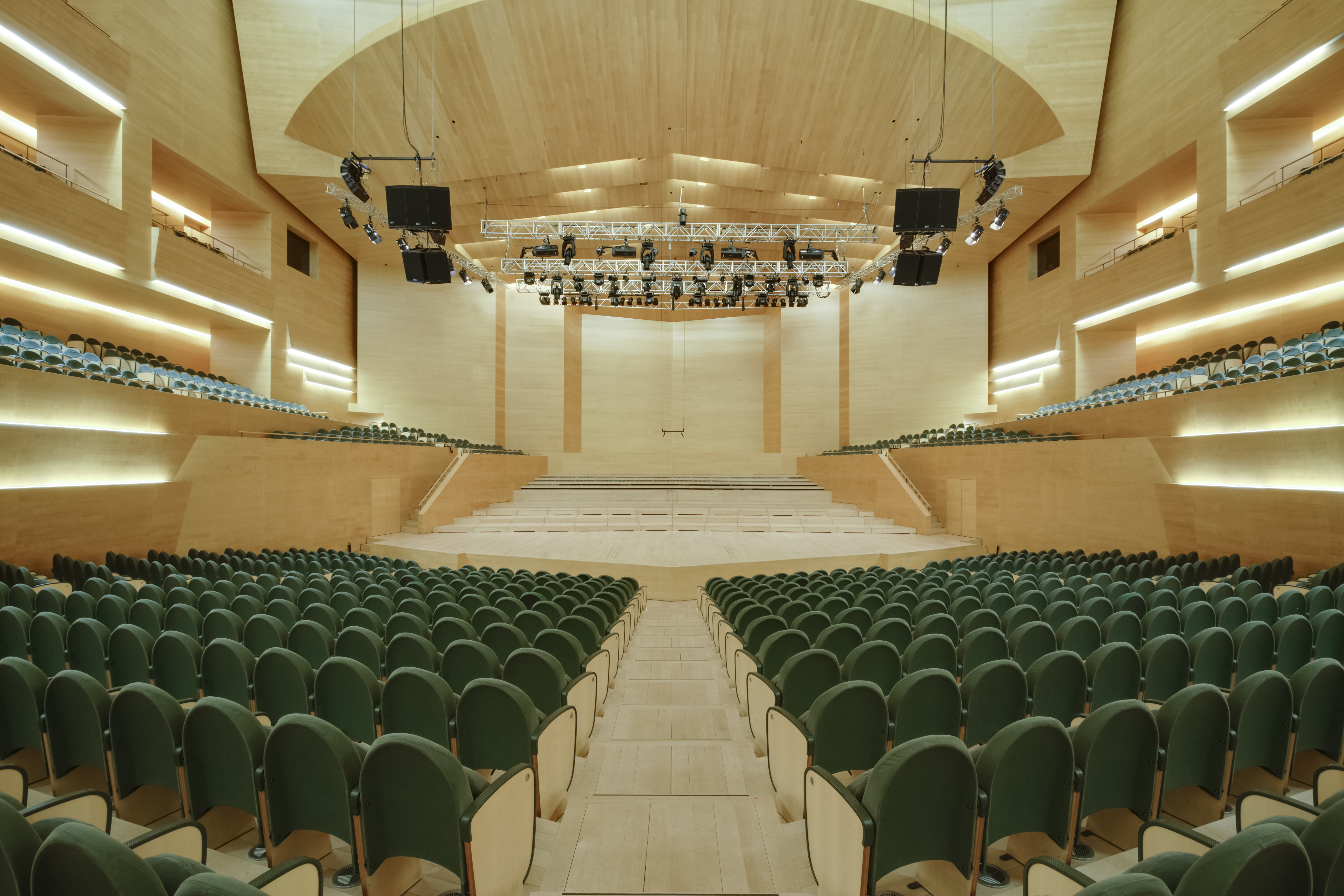 A modern empty auditorium with rows of green seats arranged on a slope towards a wide stage. The space is made of light wood, with curved walls and ceiling that add warmth. Above the stage there is a suspended lighting and sound structure, and on the sides there are tiered seating levels with integrated lighting.