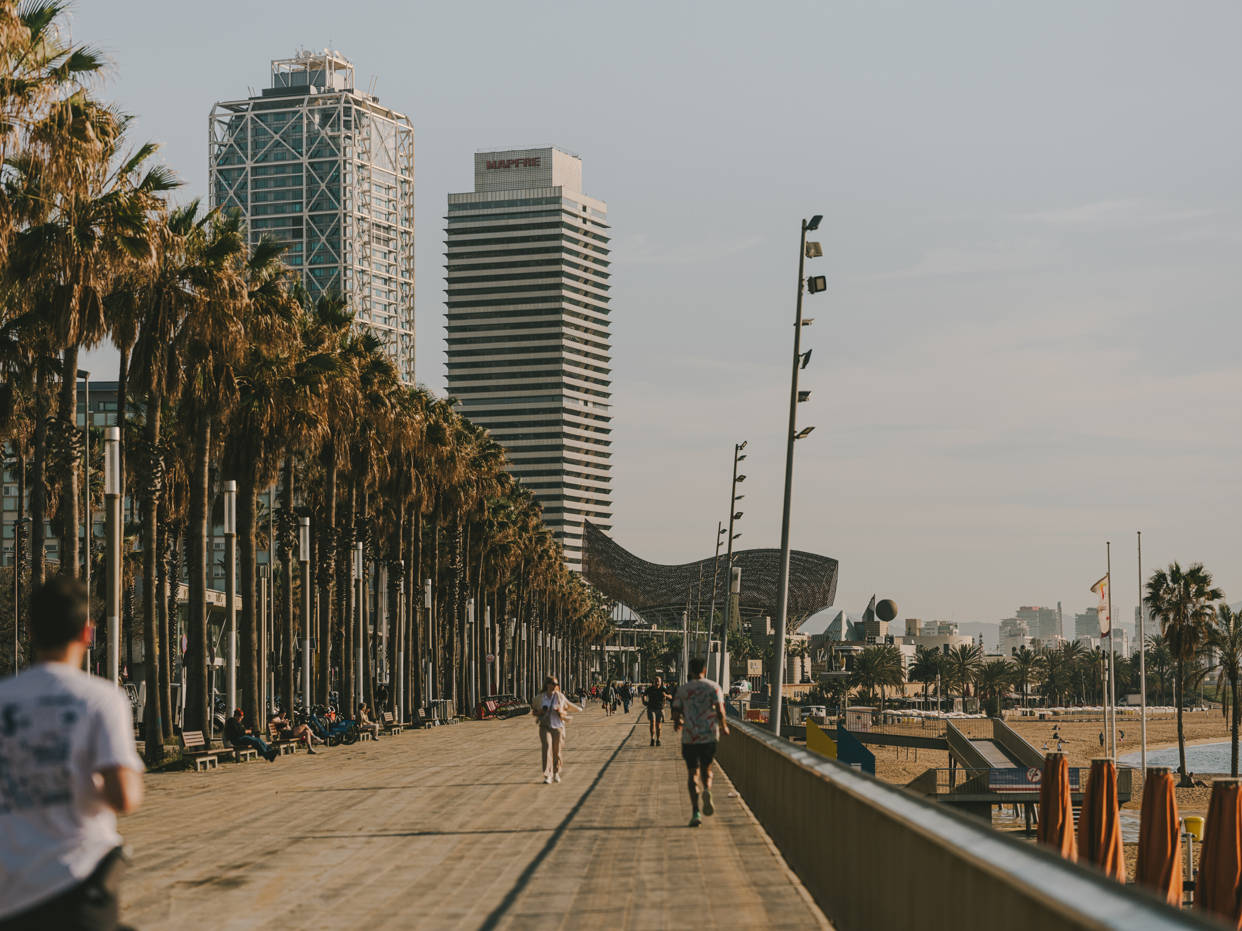 Barcelona seafront promenade with palm trees, pedestrians and runners, with the Port Olímpic towers and the beach in the background under a clear sky.