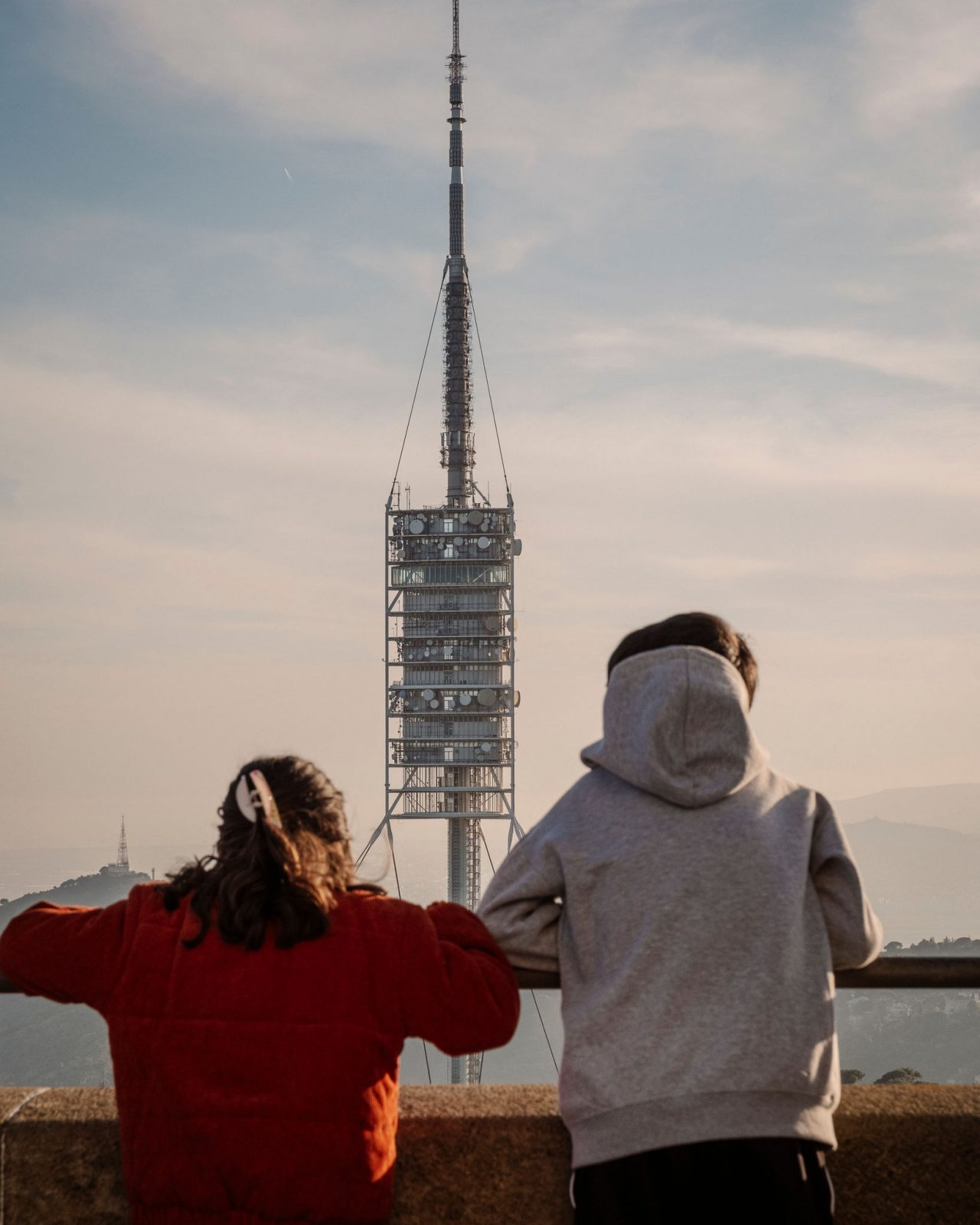 Dos personas de espaldas observando la torre de comunicaciones de Collserola desde un mirador de Barcelona, con el paisaje urbano y las montañas al fondo.