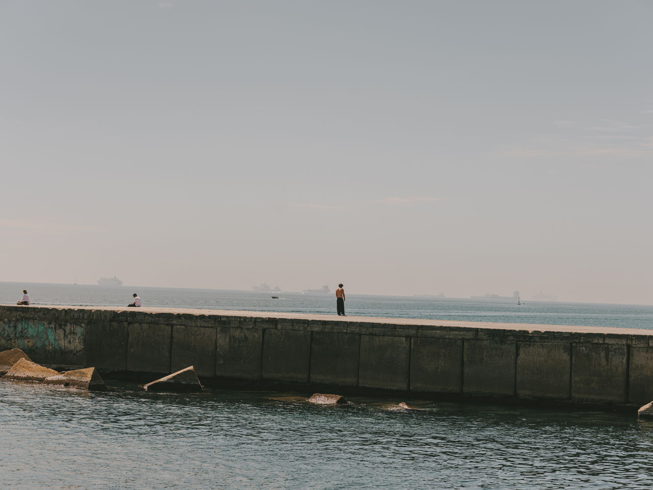 Personnes se promenant et assises sur une jetée face à la mer de Barcelone, avec des bateaux à l’horizon et des eaux calmes au premier plan.