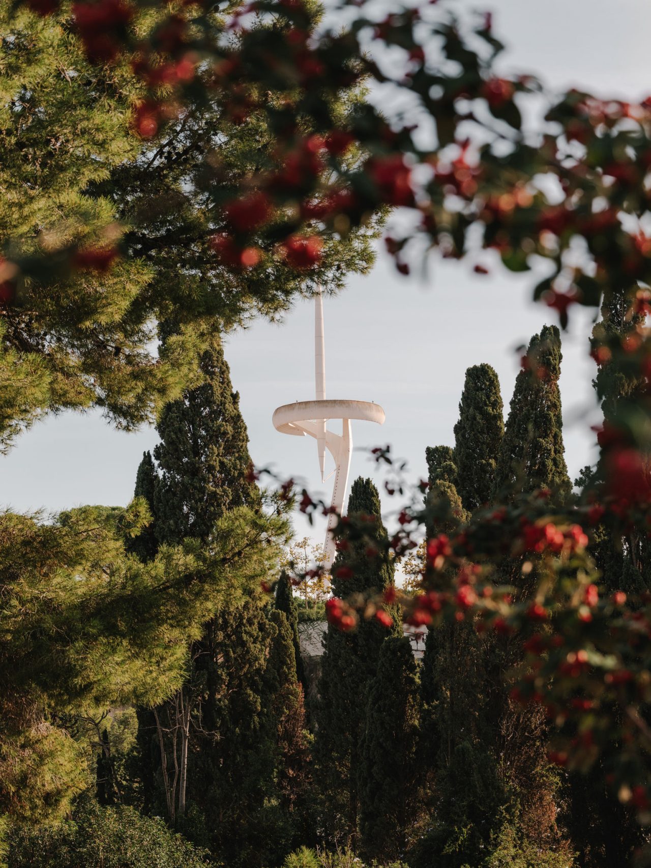 Tour de télécommunications de Montjuïc entourée de végétation, vue entre les arbres et des fleurs rouges floues au premier plan.