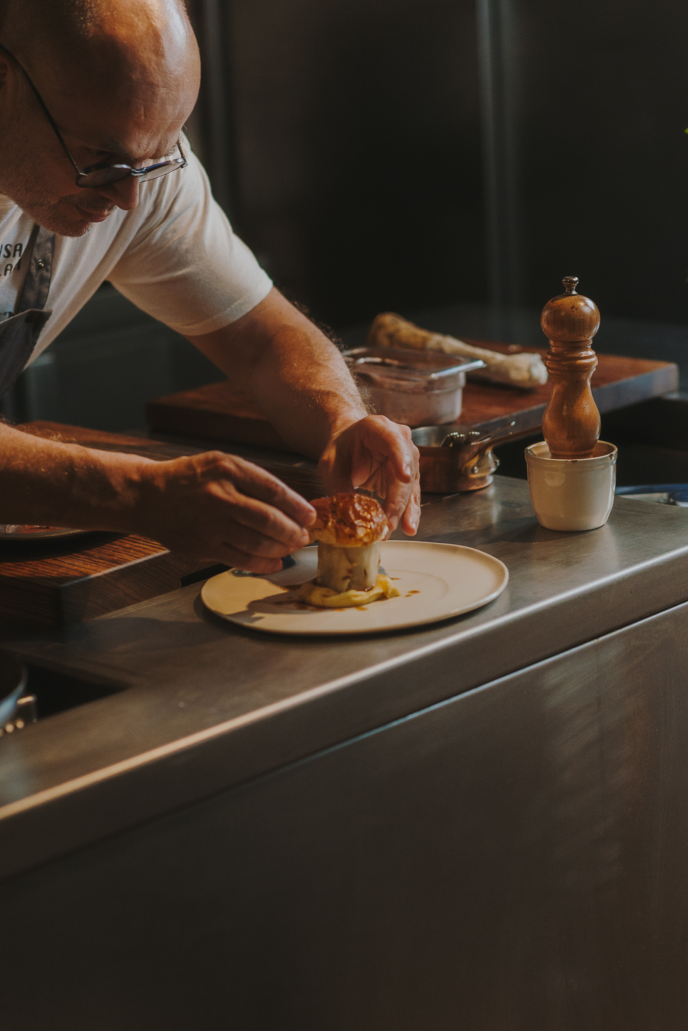 Chef, en una cocina profesional, terminando de emplatar un plato con un hongo en el centro. La mesa en la que trabaja es de acero inoxidable. Delante del chef hay tablas de cortar de madera y un molinillo de pimienta. Las manos del chef están en el centro de la imagen, prestando atención a la presentación precisa de la comida.