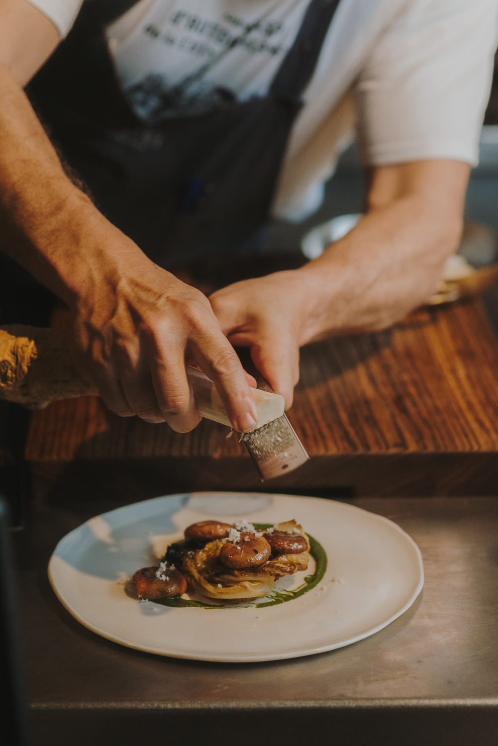 Manos de un chef decorando el plato rallando queso. Entre el chef y el plato hay una tabla de cortar de madera.