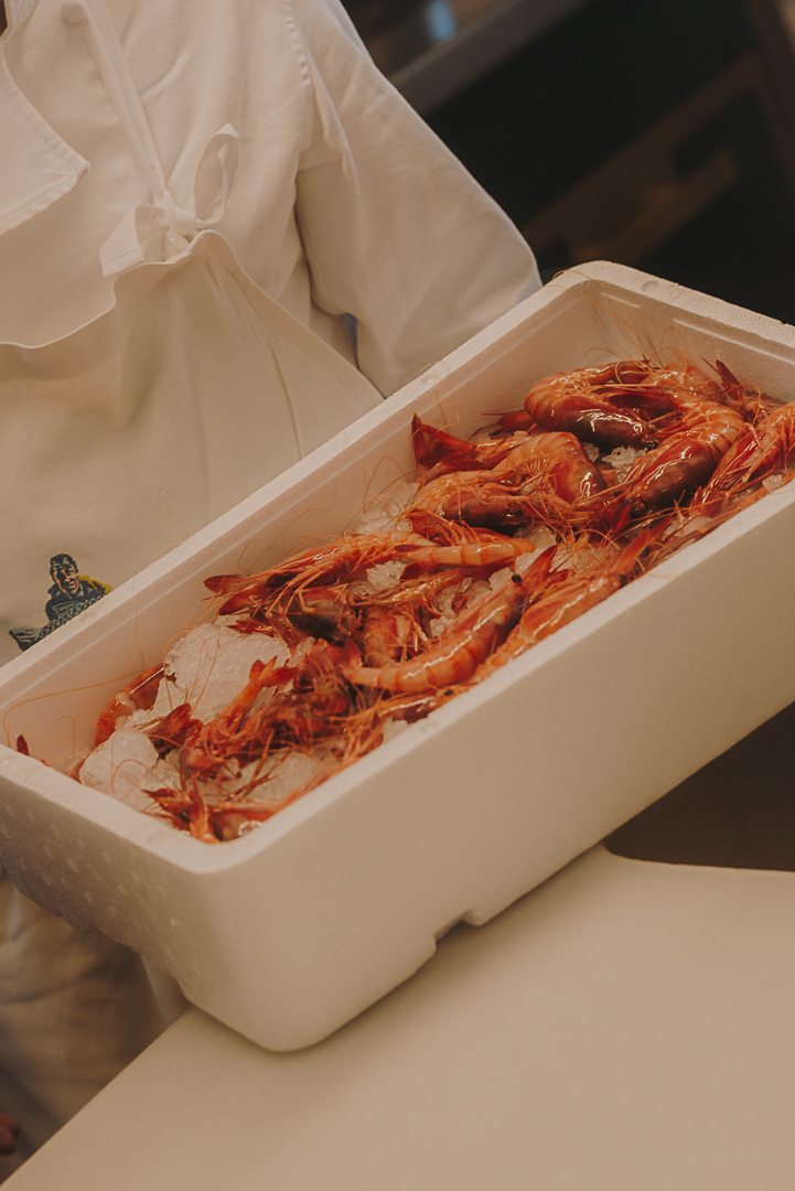 Persona sosteniendo una caja blanca de porexpan con varias gambas rojas frescas dispuestas sobre hielo. La imagen muestra un primer plano del recipiente en un entorno de preparación gastronómica.