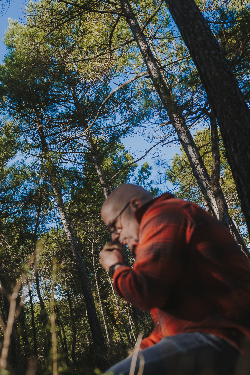 Homme portant une chemise à carreaux rouge dans une forêt, entouré de grands arbres. Il sent un champignon qu’il tient entre ses doigts.
