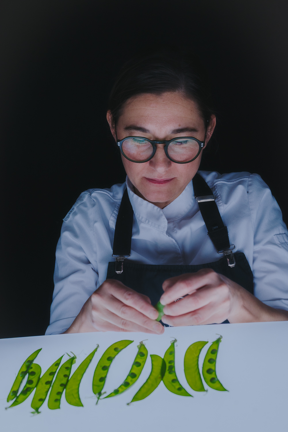 Chef portant des lunettes et un uniforme de cuisine manipulant des petits pois verts sur une table éclairée, sur fond noir.
