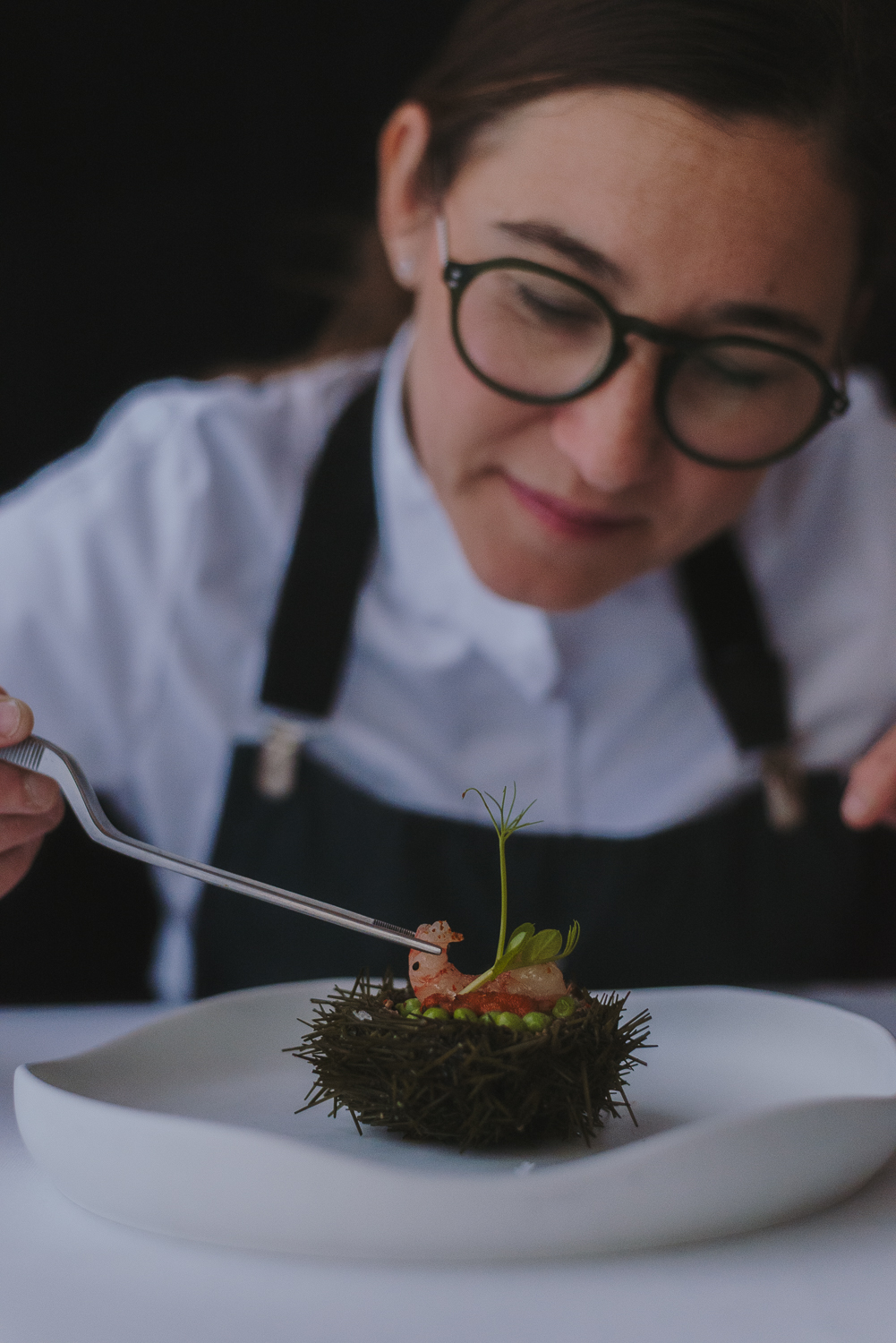 Chef portant des lunettes décorant un plat gastronomique avec des petits pois et des pousses vertes sur une base sombre, dans une présentation soignée.