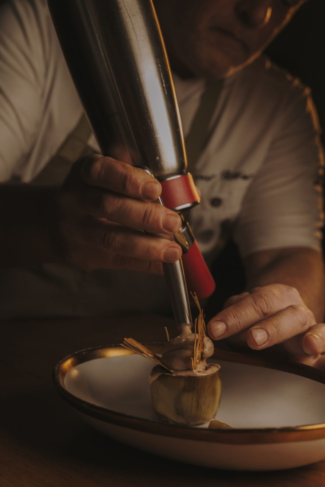 Mains d’un chef utilisant un siphon culinaire pour terminer un plat d’artichaut sur une assiette décorative. Le chef porte un t-shirt à manches courtes et un tablier gris. La lumière est chaude.