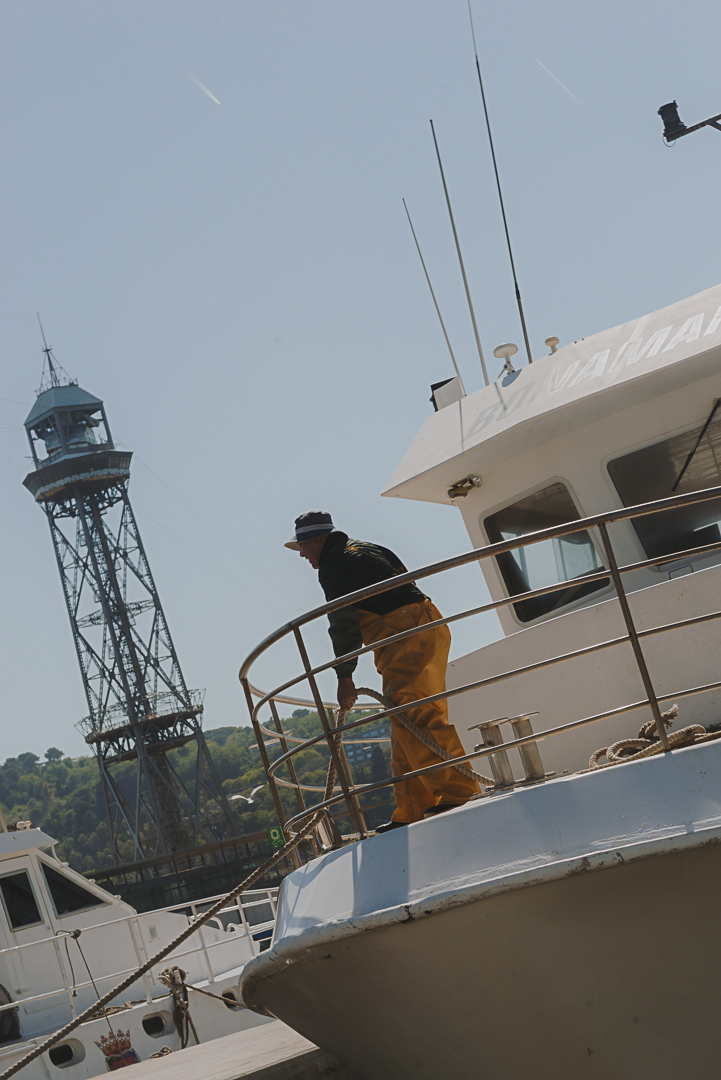 Homme travaillant sur le pont d’un bateau au port de Barcelone, avec la tour du téléphérique du port en arrière-plan. Il porte des vêtements de travail orange et noirs et est appuyé sur la rambarde du bateau par une journée ensoleillée.