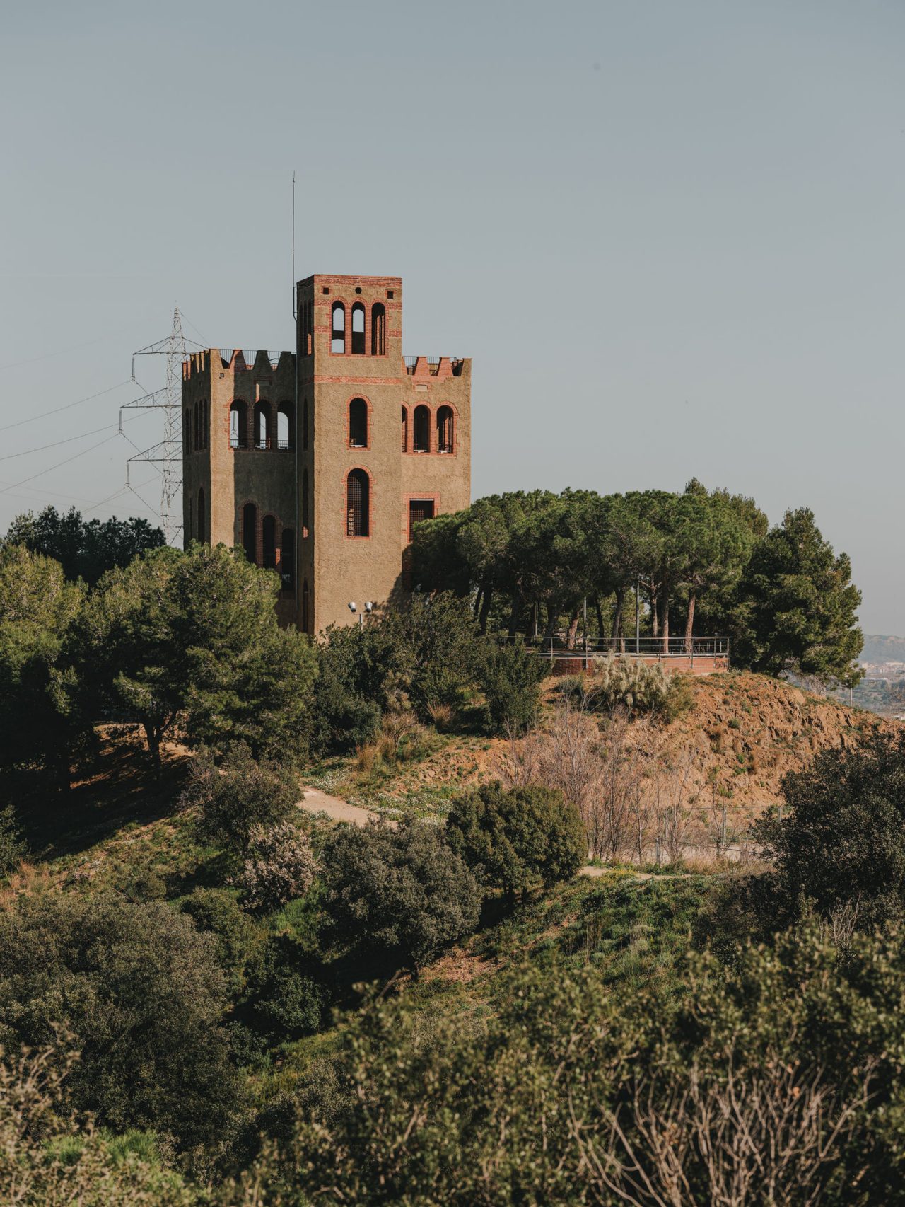 Tour de guet en briques située sur une colline de Collserola, entourée de pins et de végétation méditerranéenne, avec une ligne électrique en arrière-plan.