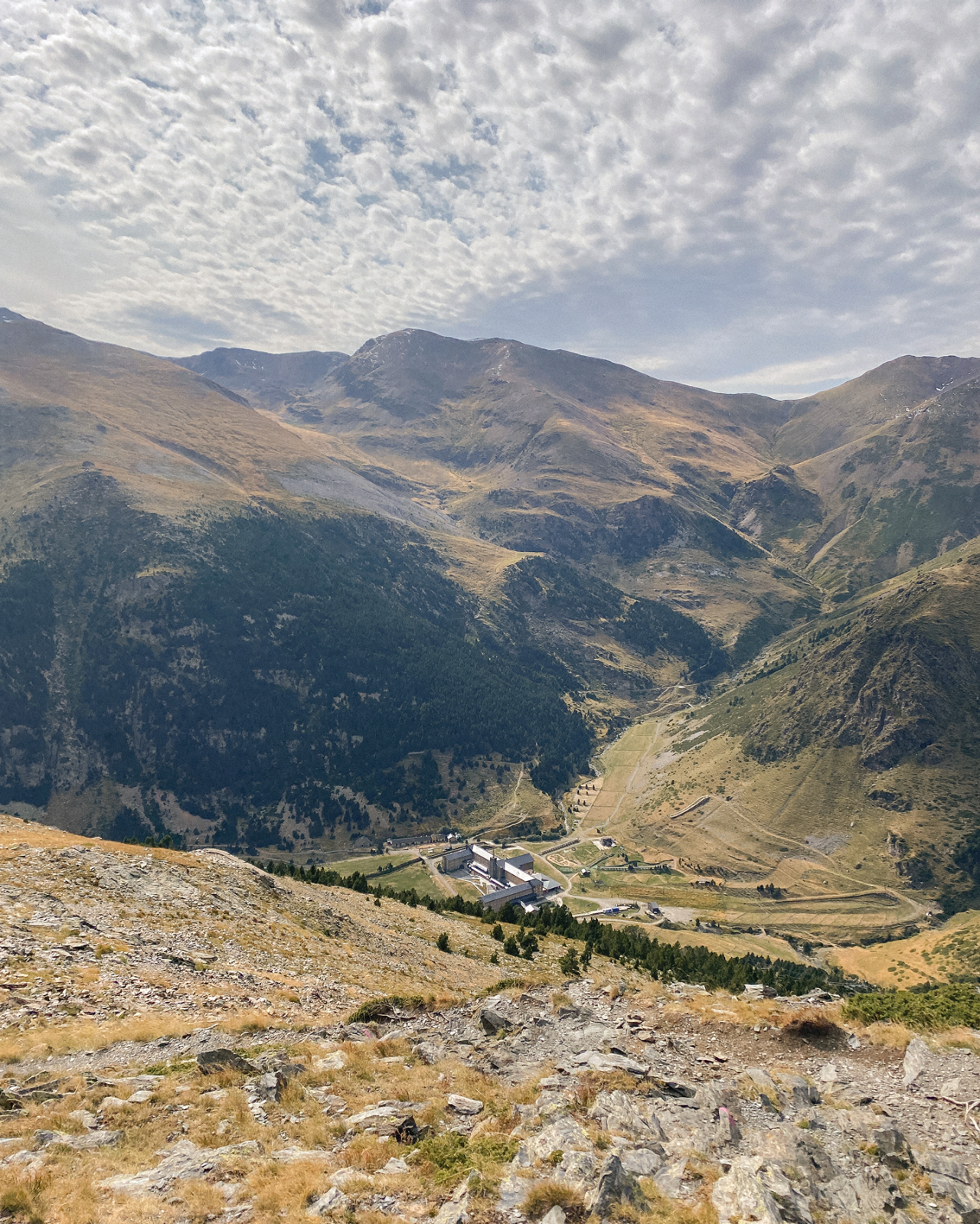 Vue panoramique d’une vallée de montagne avec des chemins, des bâtiments et un relief ondulé sous un ciel nuageux.