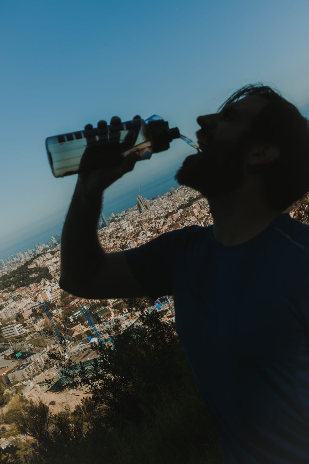 Homme buvant de l’eau d’une bouteille dans une zone surélevée, avec une vue panoramique de Barcelone et de la mer en arrière-plan.