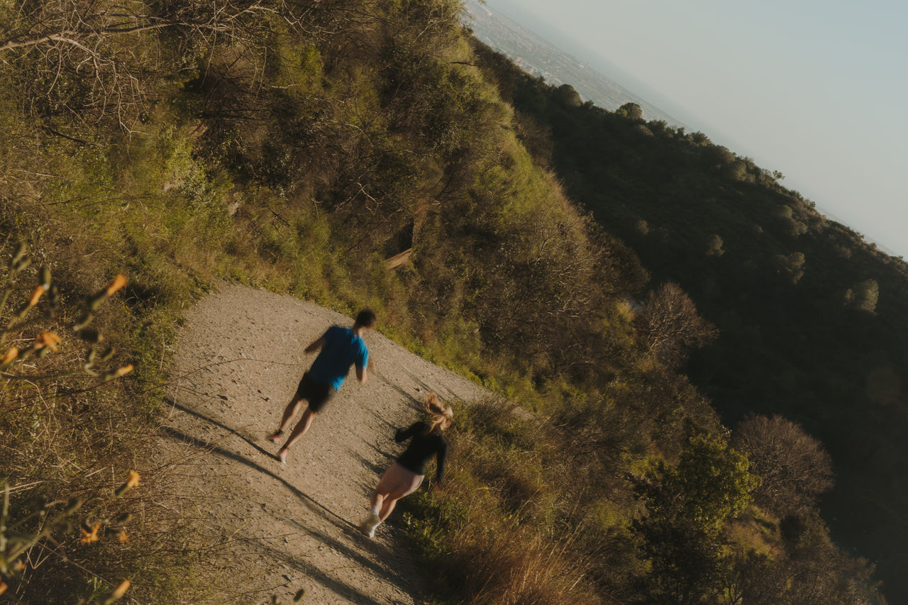Deux personnes courent sur un chemin de montagne entouré de végétation, avec le versant de Collserola en arrière-plan.