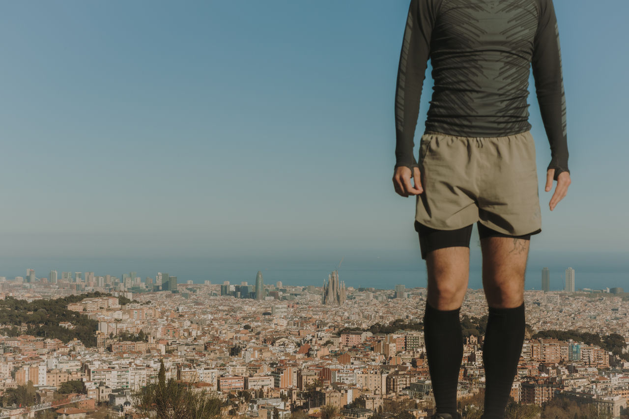 Man standing in an elevated mountain area, with a panoramic view of Barcelona, the Sagrada Família and the sea in the background.