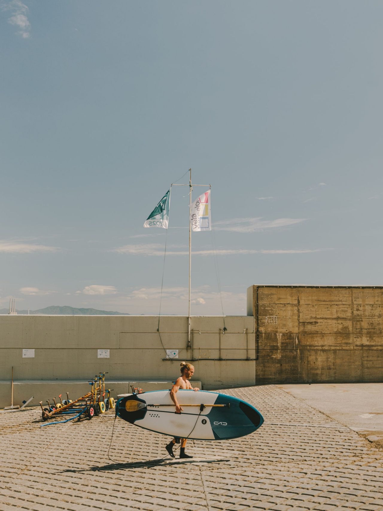 Persona che trasporta una tavola da paddle surf in un’area pavimentata vicino al mare, con bandiere, pagaie e l’orizzonte marittimo sullo sfondo.