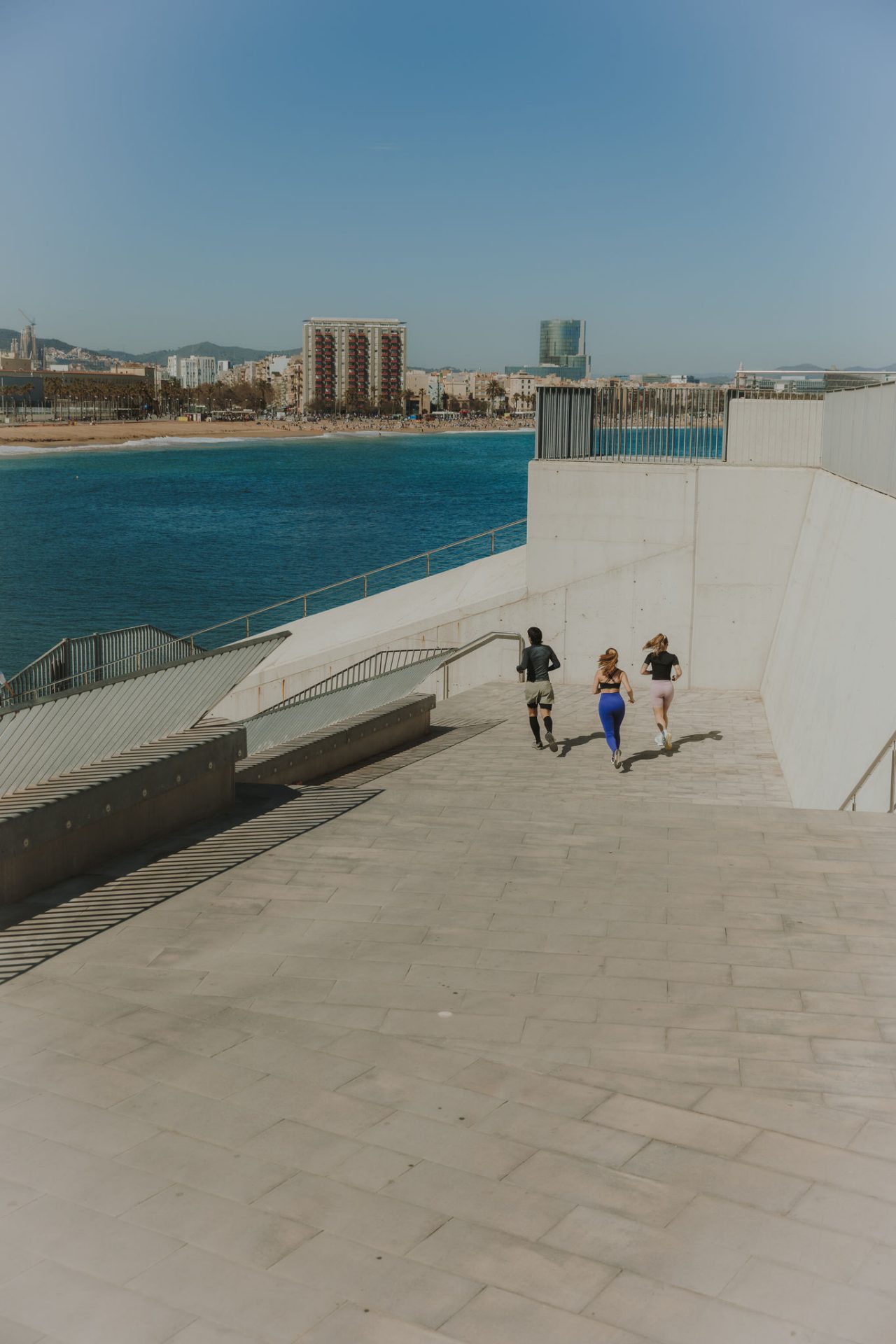 Tre persone corrono lungo una scalinata accanto al mare, con la spiaggia e gli edifici di Barcellona sullo sfondo.